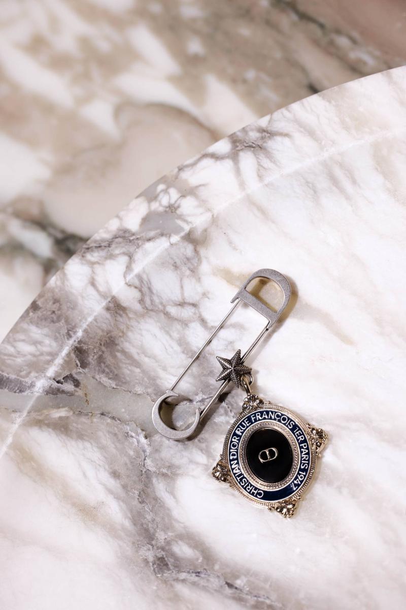 مشبك للزينة من Dior Men تميّزه ميدالية تحمل شعار ديور ونقشًا باسم المؤسس وعنوان الدار في باريس وتاريخ تأسيسها. 