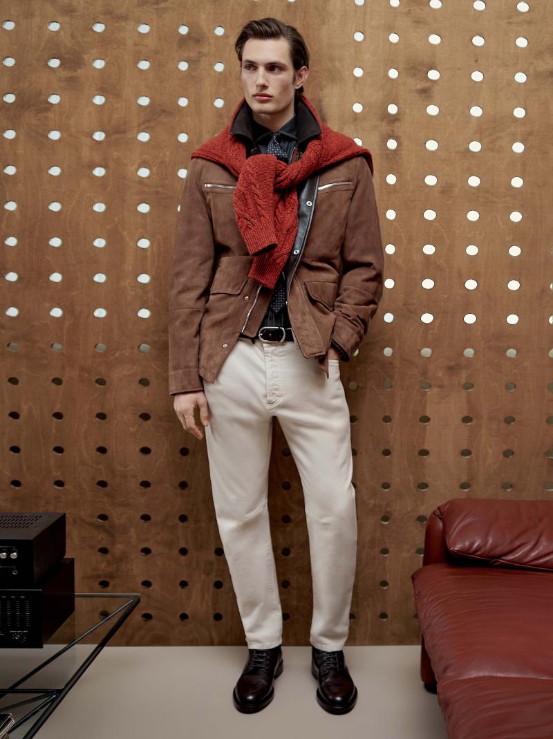 كنزة باللون الأحمر محبوكة من الكشمير، وسترة من الجلد المقلوب، وقميص من نسيج الدنيم، وسروال مشغول من الدنيم، وربطة عنق من الحرير تزدان بنقوش الجاكارد، وحزام جلدي مضفّر، وحذاء ديربي مشغول من جلد العجول. 